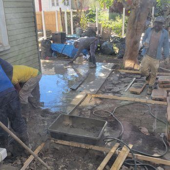 Construction workers laying stone patio in a backyard. Wet cement and wooden frames are visible.