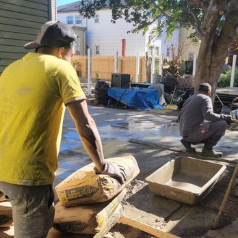 Two workers in a backyard, one holding cement bags, the other squatting by a mortar tub. Sunny day.