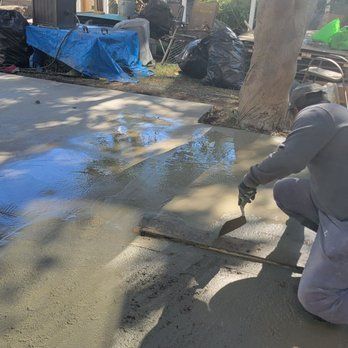 Person smoothing wet concrete with a trowel outdoors; blue tarp, black trash bags, and tree in the background.