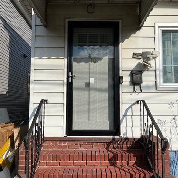 Brick steps leading to a house with a black-framed screen door and white siding. Black railing on each side.