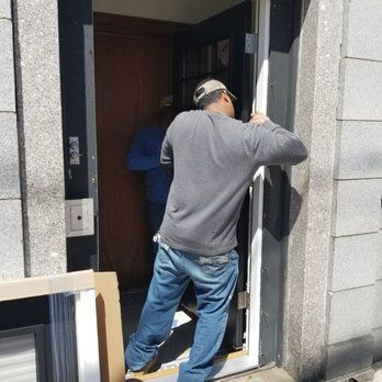 Person adjusting a door frame. Another person stands inside the doorway. Exterior view, bright daylight.