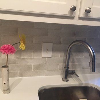 Kitchen sink with faucet, white countertops, gray tile backsplash, and flowers in a birch vase.