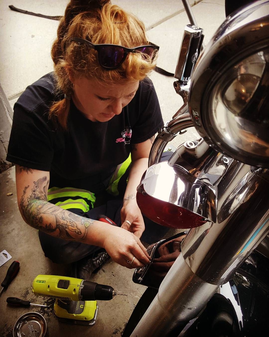 Teresa Ley with tattoos fixing a motorcycle, wearing sunglasses, kneeling outdoors.