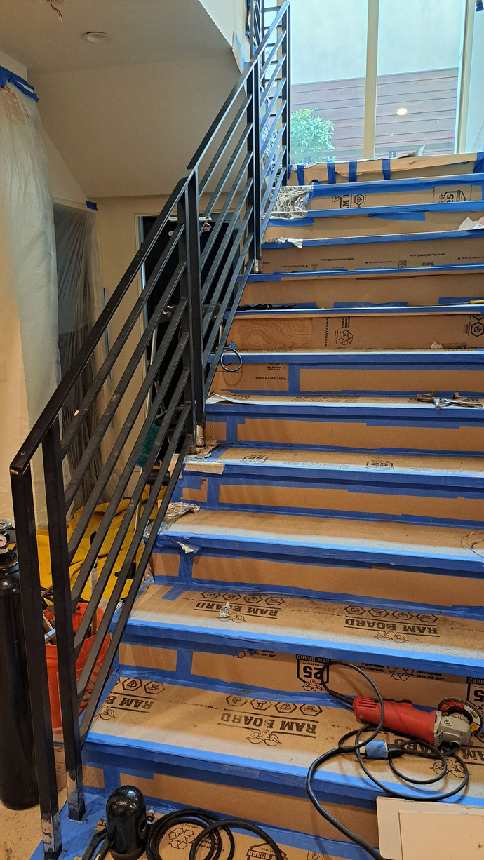 Staircase with horizontal black metal handrail. Steps covered in brown paper, blue tape. Grinder tool rests on step.