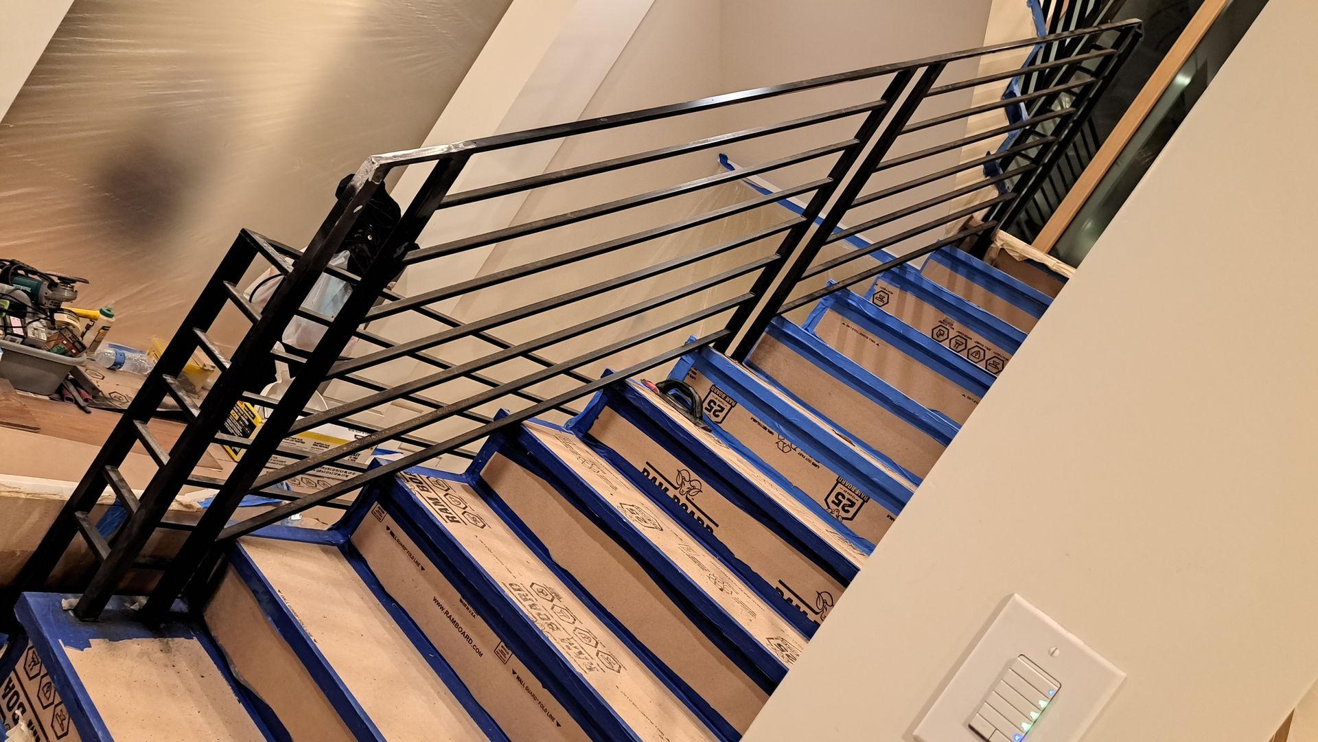 Staircase with black metal railing. Stairs are covered in blue tape and cardboard. Beige walls.