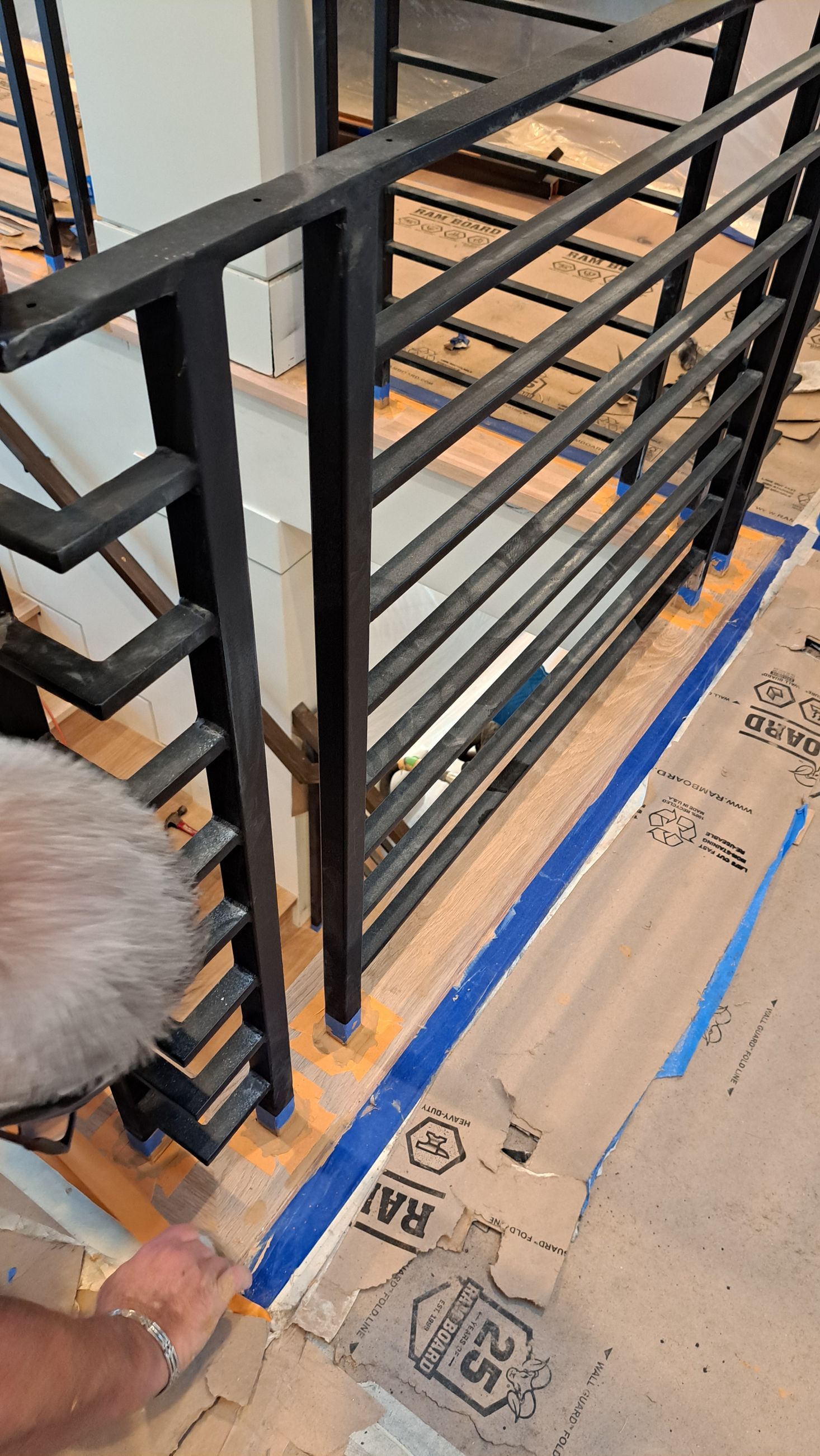 Person taping around a black metal railing on a wood surface. Blue tape marks the perimeter.