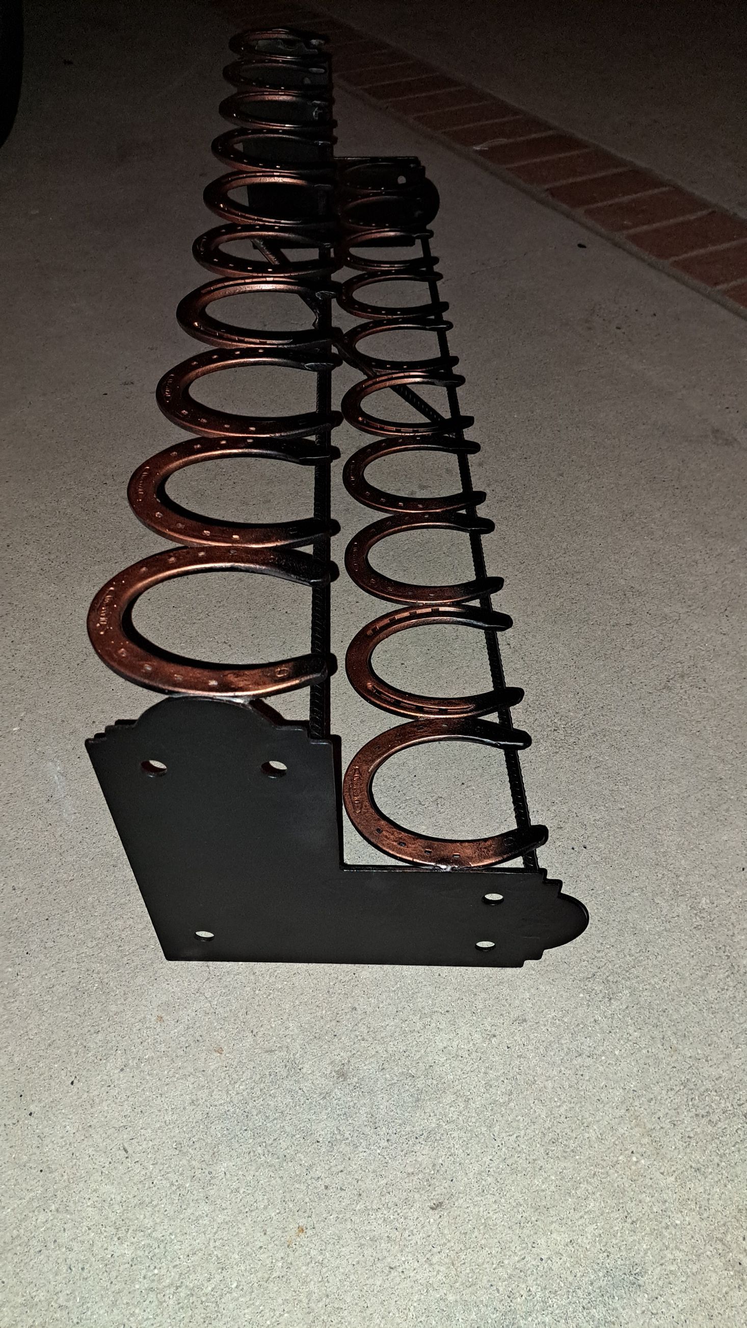 A black metal horseshoe rack with copper-colored horseshoes. It's angled against a concrete surface.