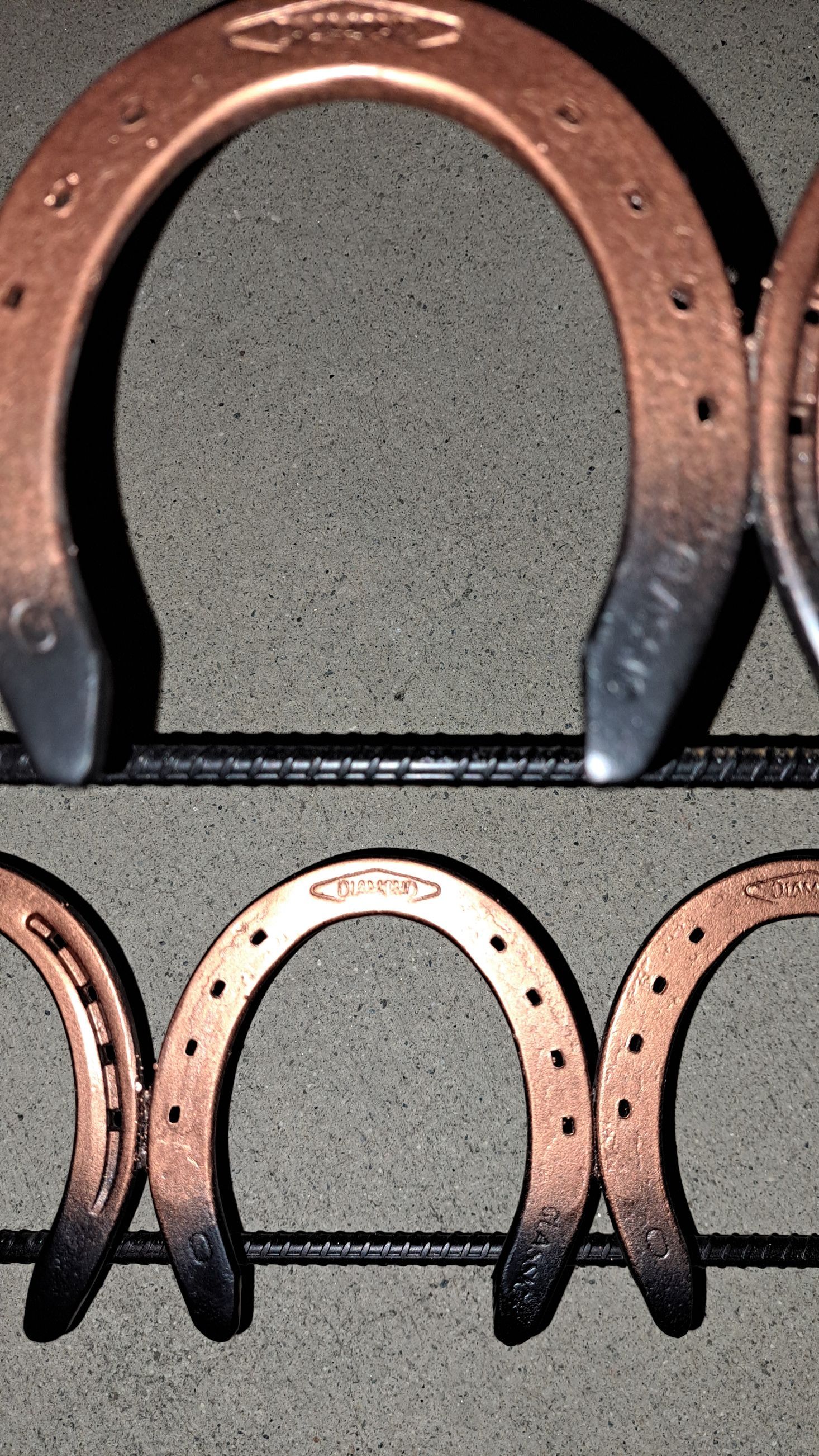 Copper-colored horseshoes with black tips arranged in a row. They are mounted on a dark horizontal metal support.