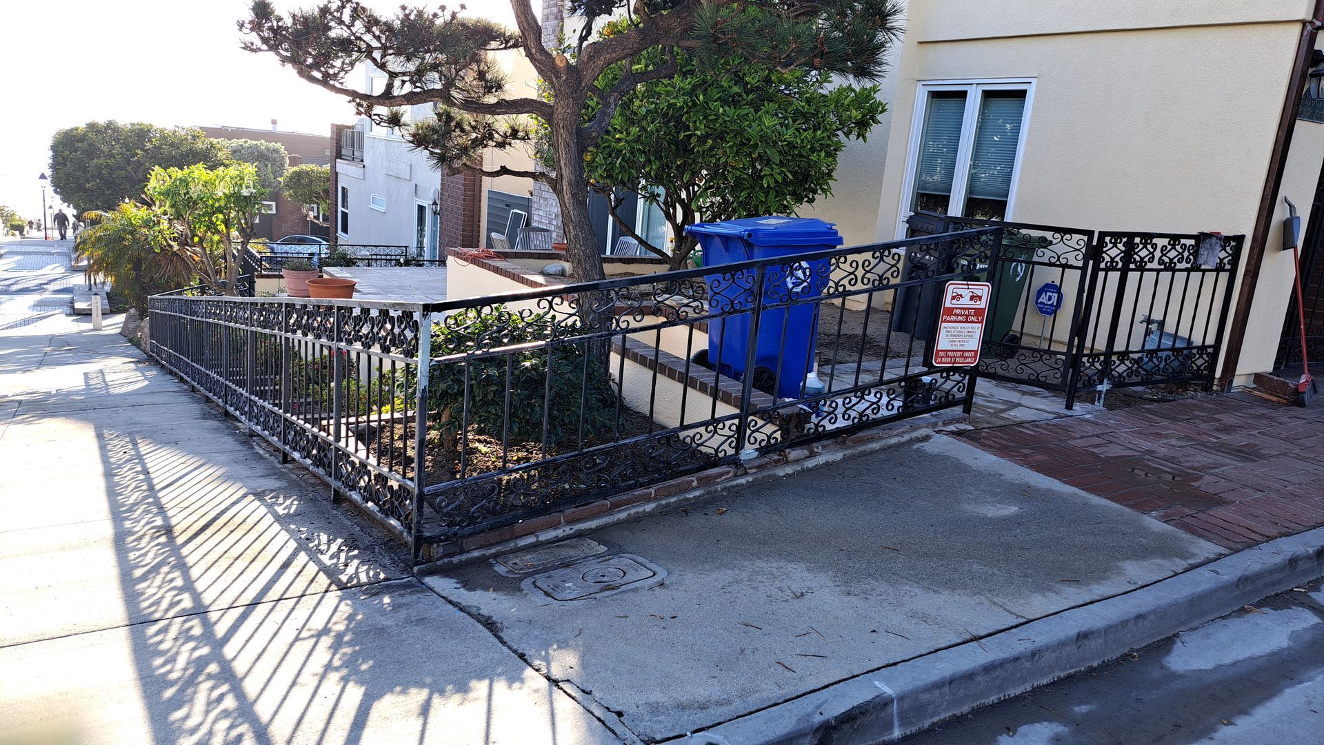 Black wrought iron fence borders a landscaped area with a blue trash bin. A light-colored building is in the background.