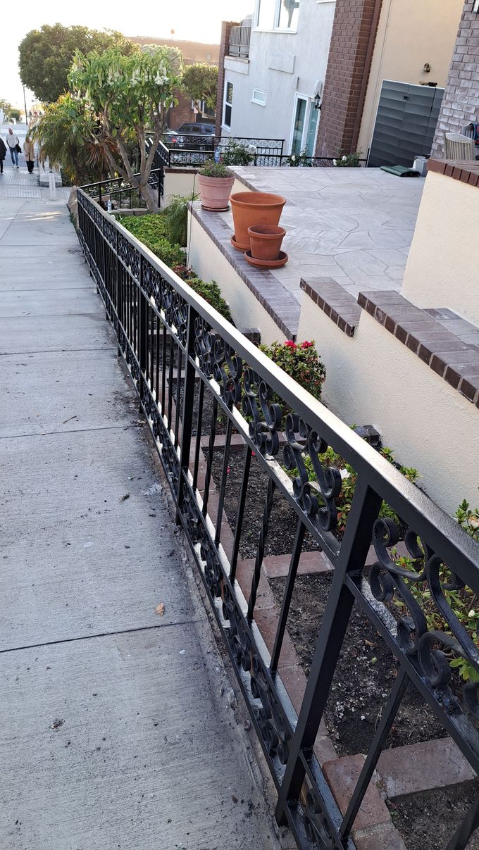 Black decorative metal fence along a sidewalk, with plants, pots, and a brick wall.