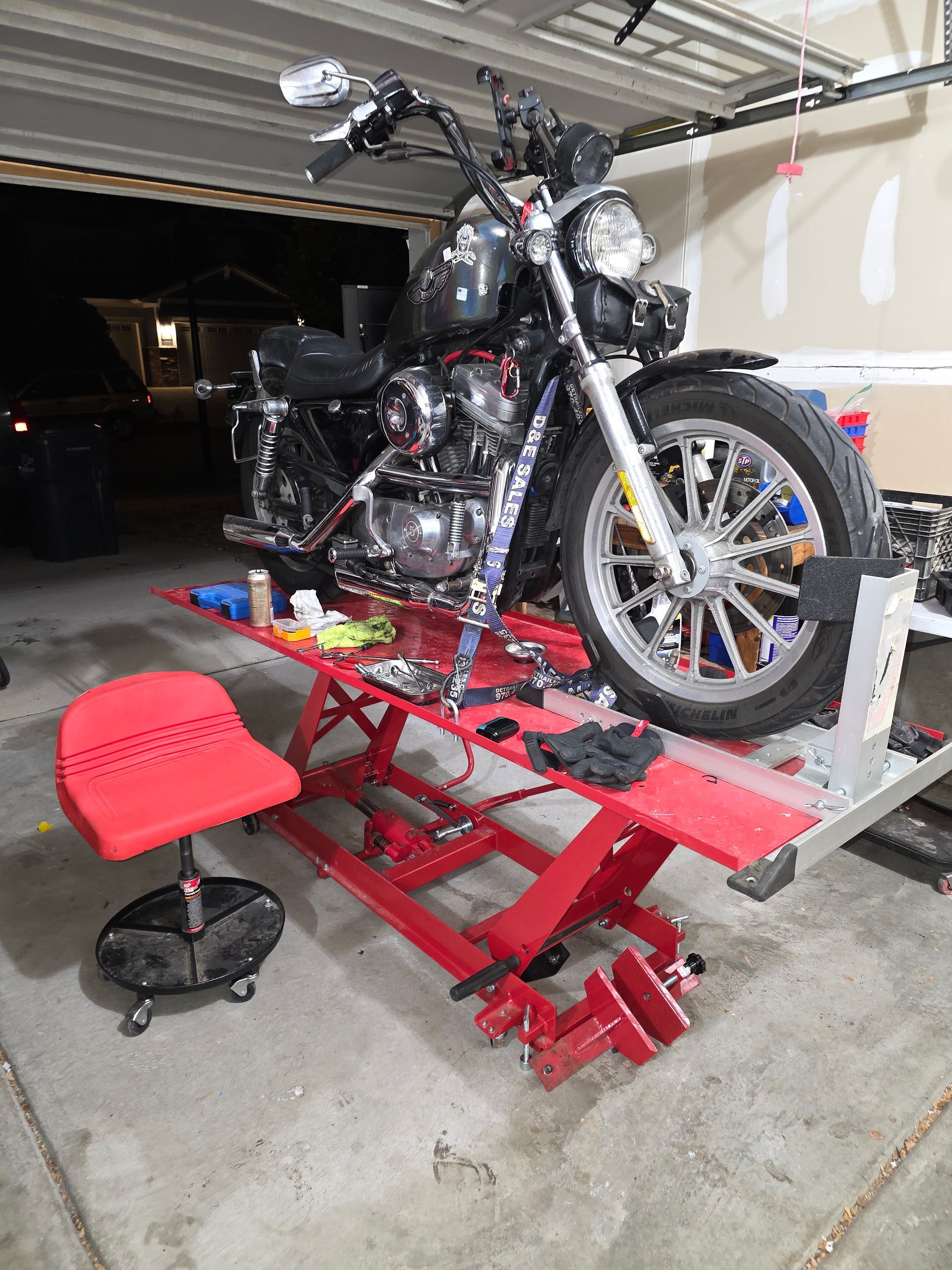 Motorcycle on red lift in garage, red rolling chair beside it, tools scattered around.