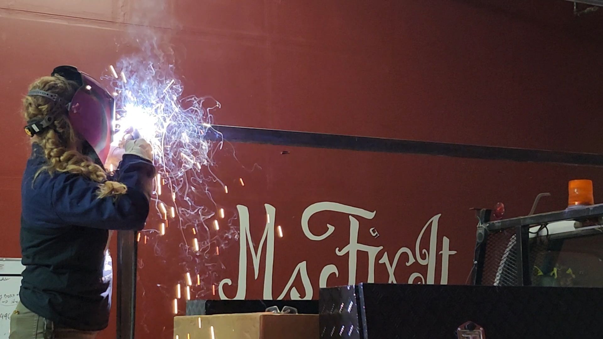Teresa Ley, Ms Fix It  welding metal bar on a red surface, sparks flying.