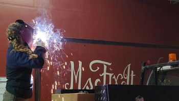 Teresa Ley, Ms Fix It welding metal bar on a red surface, sparks flying.