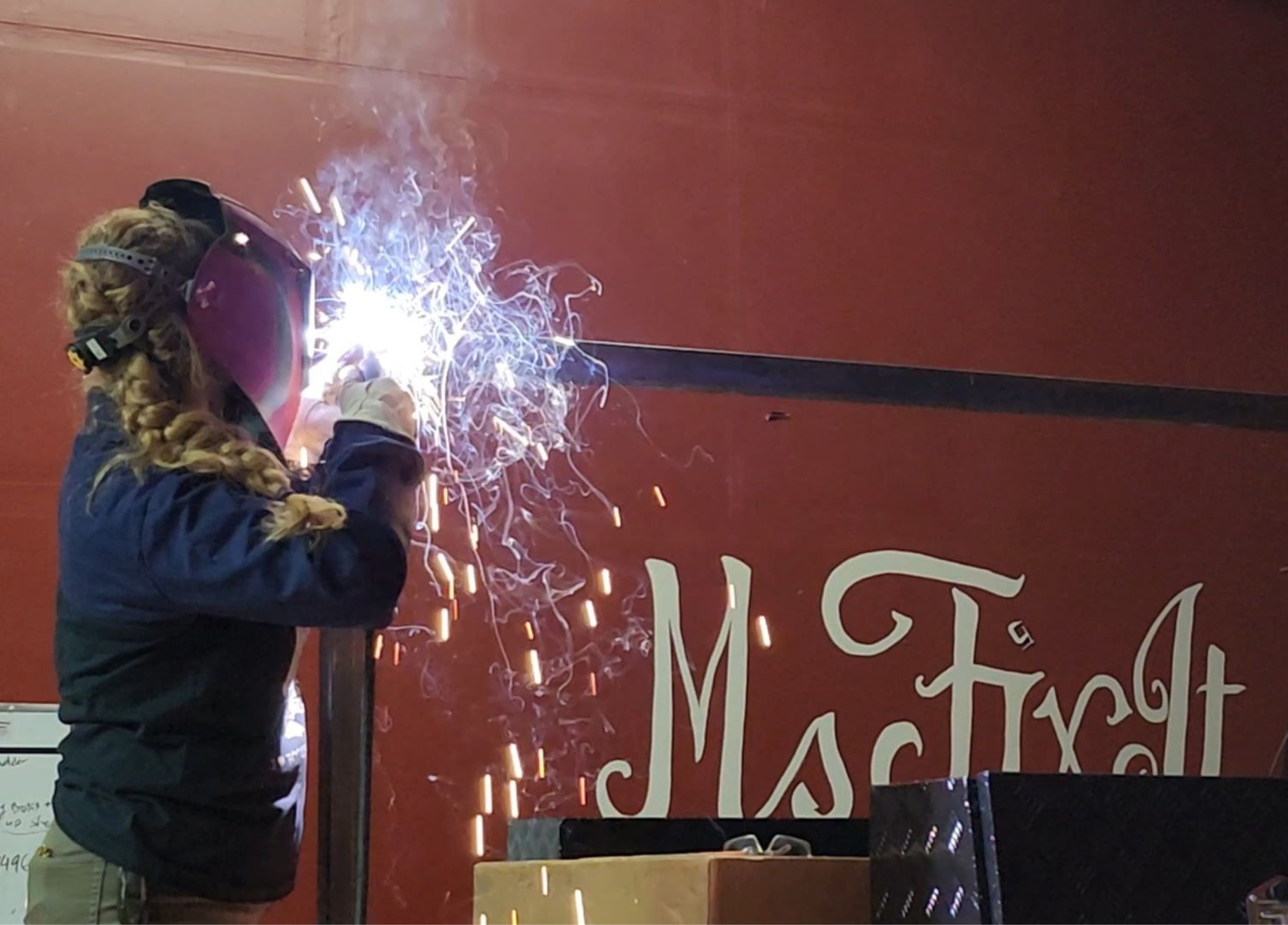 Teresa Ley Woman welding metal with sparks flying, wearing protective gear, near red wall with 