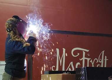 Teresa Ley Woman welding metal with sparks flying, wearing protective gear, near red wall with