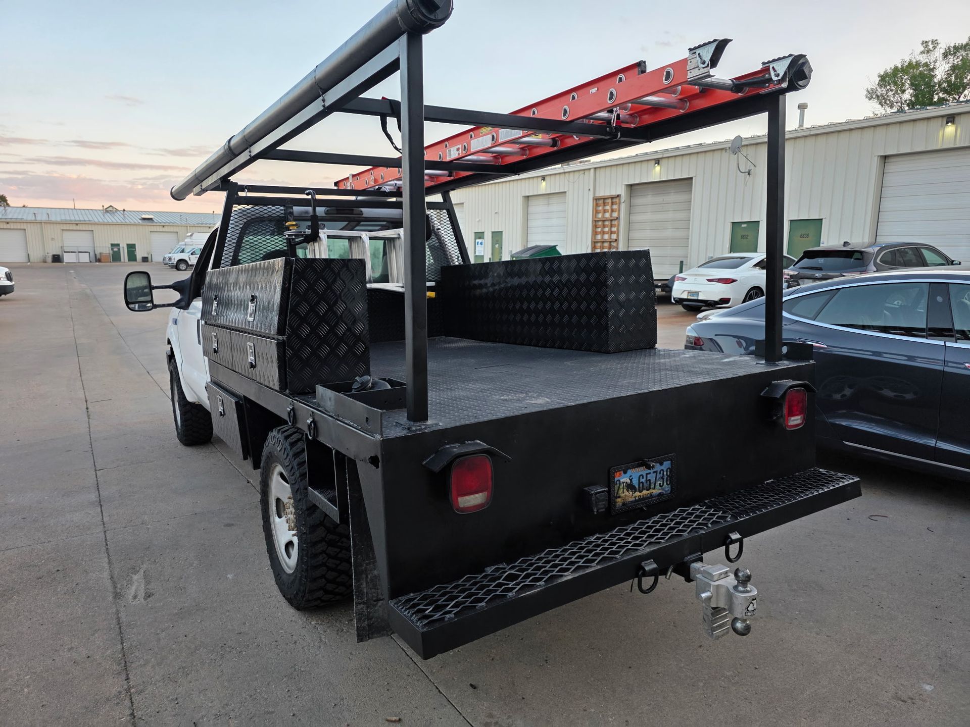 White work truck with black flatbed, ladder rack, toolboxes. Parked outdoors.