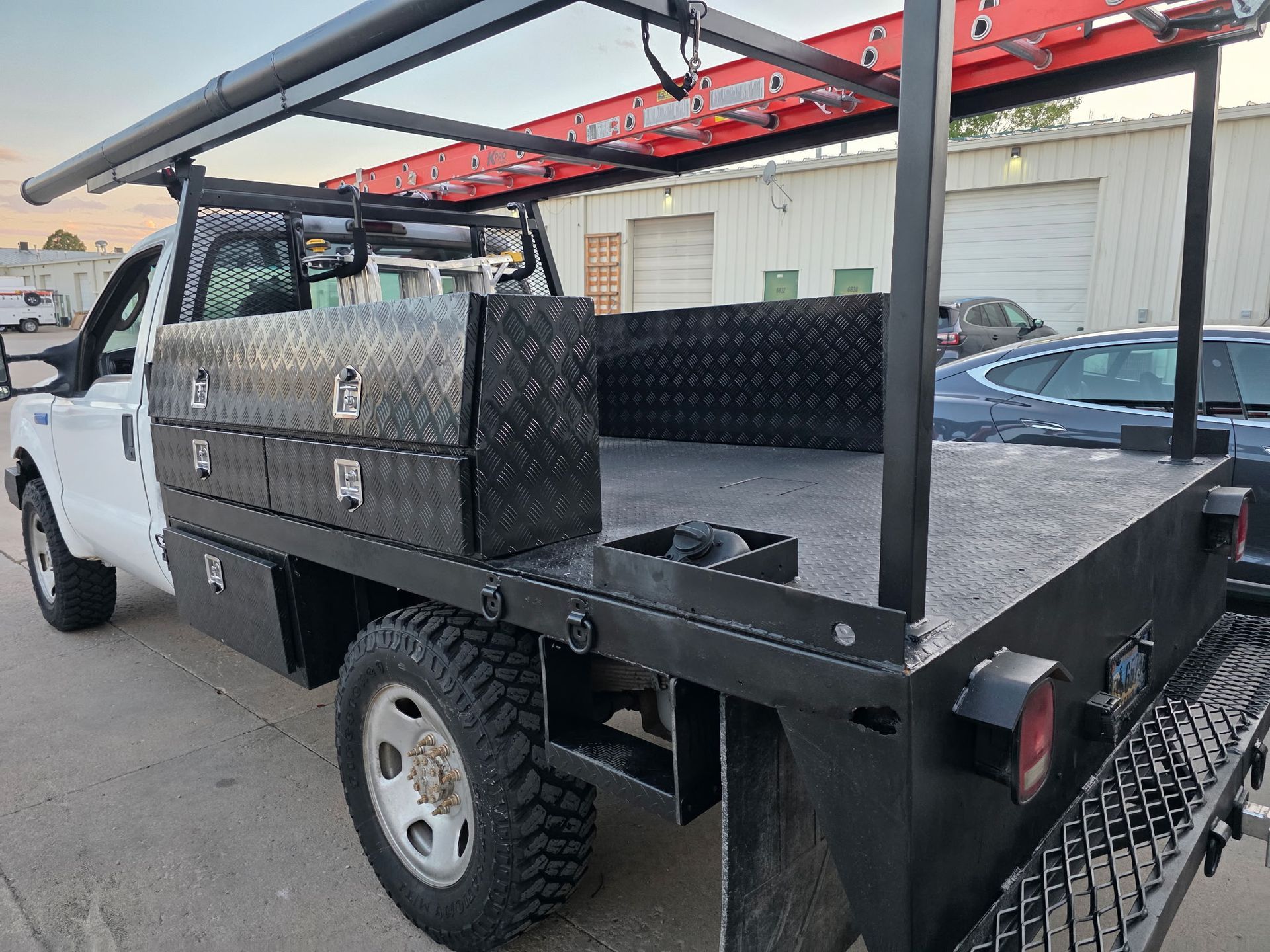 White work truck with black flatbed, toolboxes, ladder rack, and red ladder, parked outside.