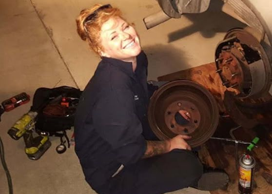 Woman smiling, holding brake drum, working on a vehicle. Tools and car parts in garage setting.