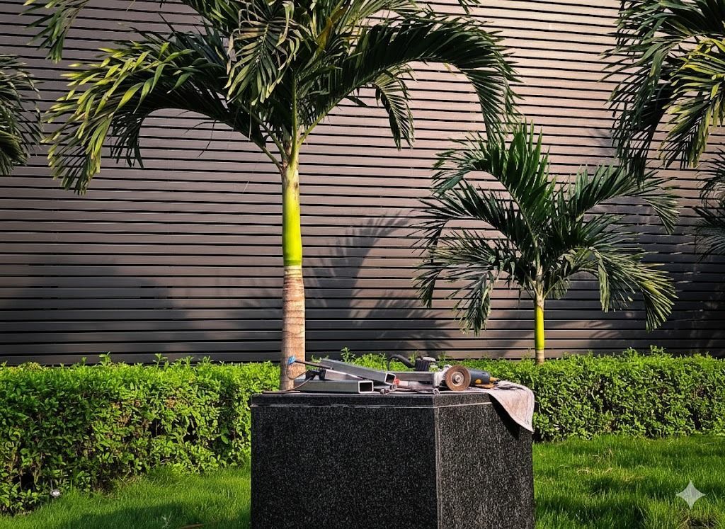 Palm trees with yellow accents stand near a dark granite planter on green grass, with a textured wall background.