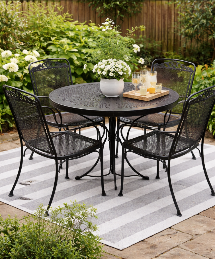 Black metal patio table and chairs on striped rug, surrounded by greenery.