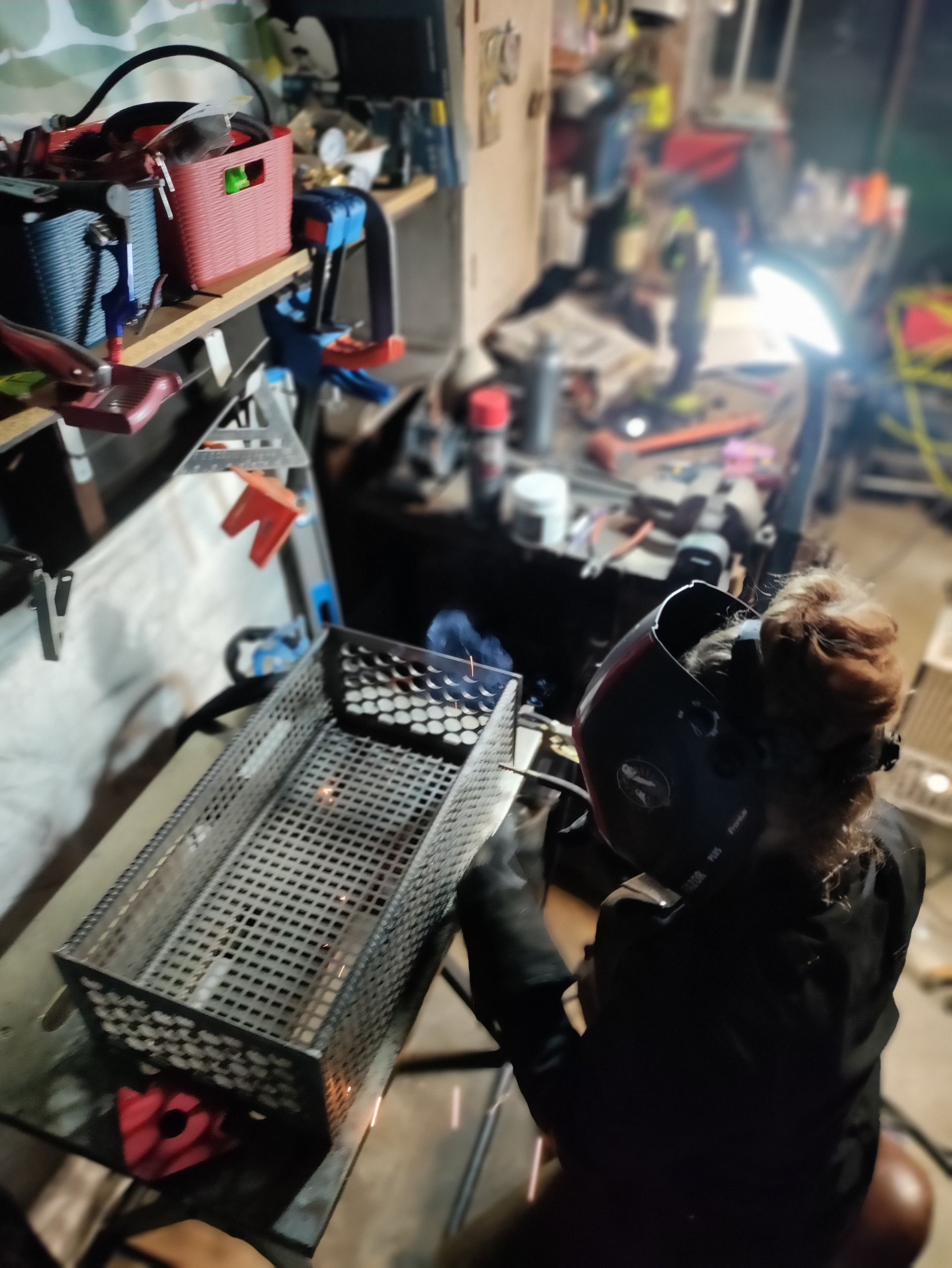 Person welding a metal basket in a workshop. Wearing a welding helmet and gloves. Tools and shelves in the background.