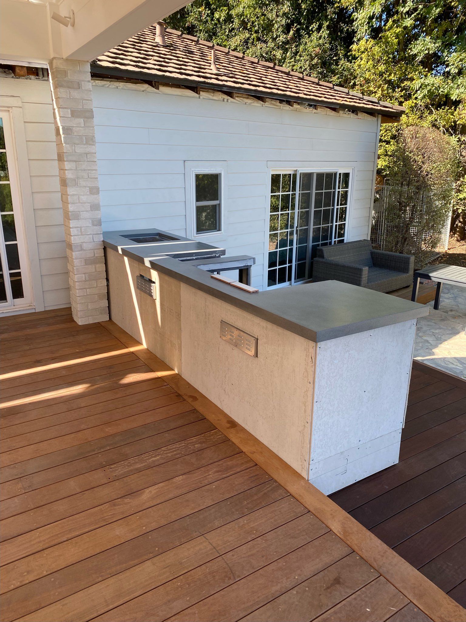 Outdoor kitchen with gray countertop, on a wood deck, with a small white building in the background.