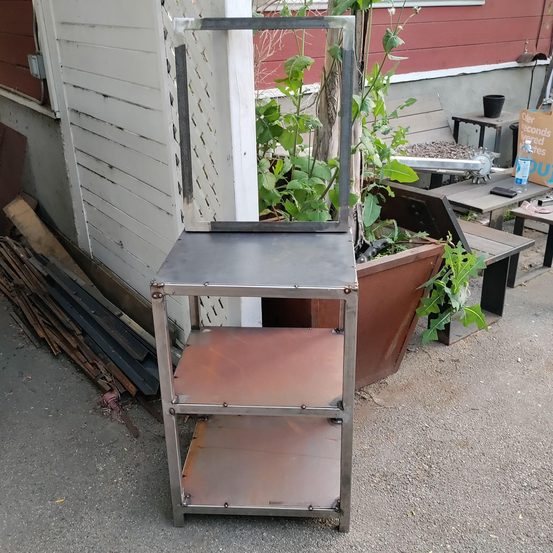 Metal cart with three shelves, a top frame, and potted plants in a backyard setting.