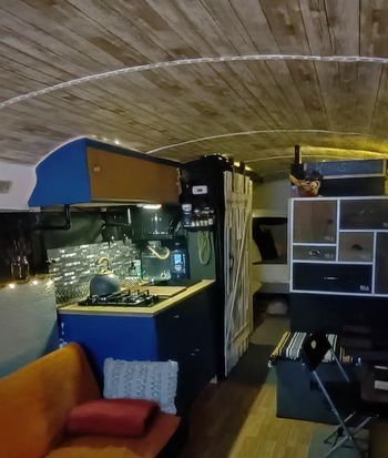 Interior of a bus converted into a living space, featuring a kitchen, seating, wooden ceiling, and storage cabinets.
