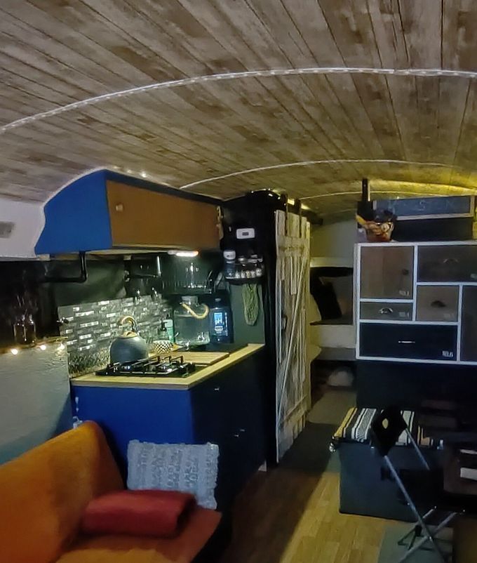Interior of a renovated bus with a kitchen, seating, and wood-paneled ceiling. Blue and black cabinets, an orange couch.