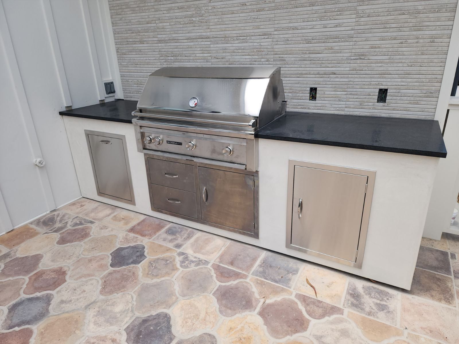 Outdoor kitchen with stainless steel grill, cabinets, and black countertop on stone patio.
