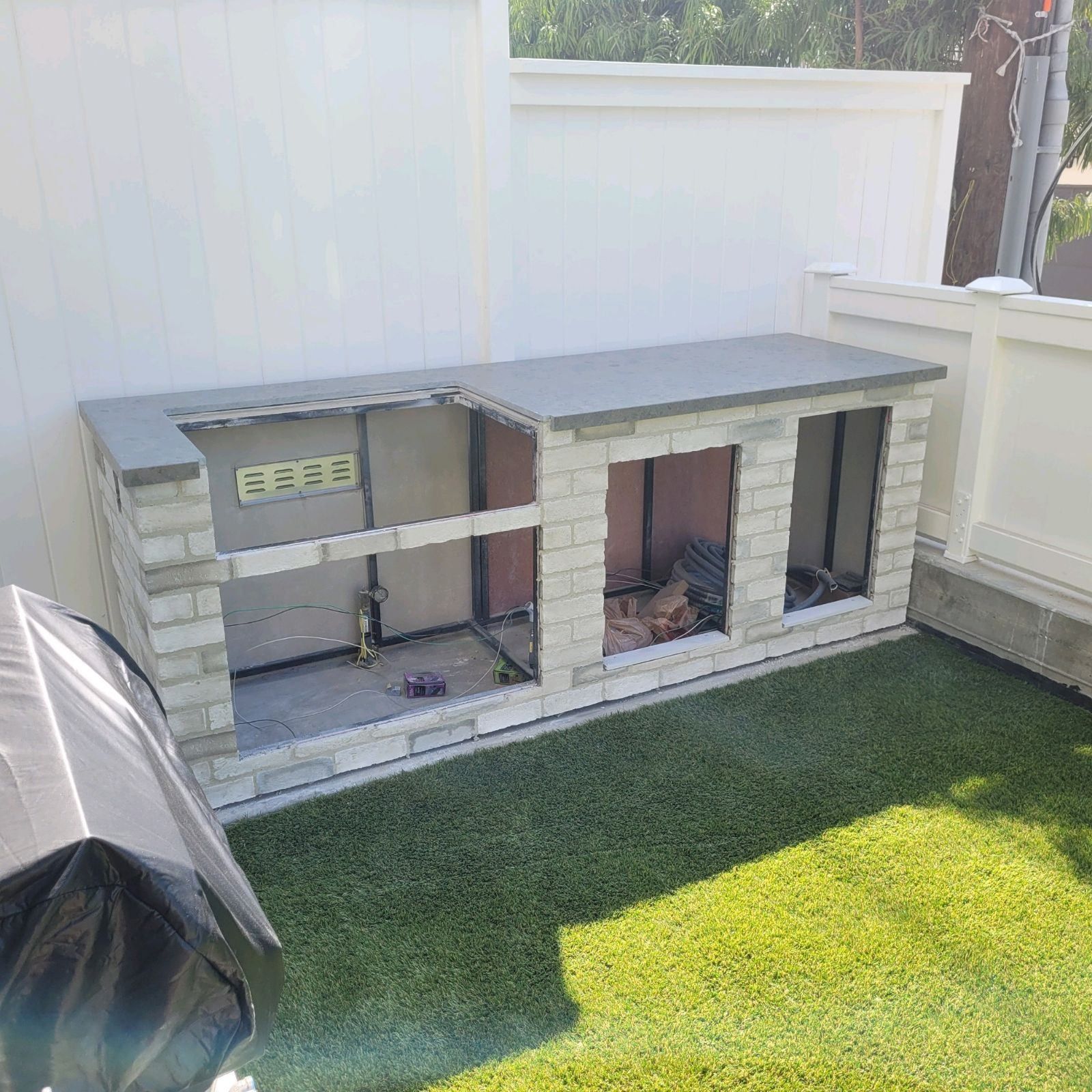 Outdoor kitchen under construction with gray stone and concrete countertop on artificial grass.