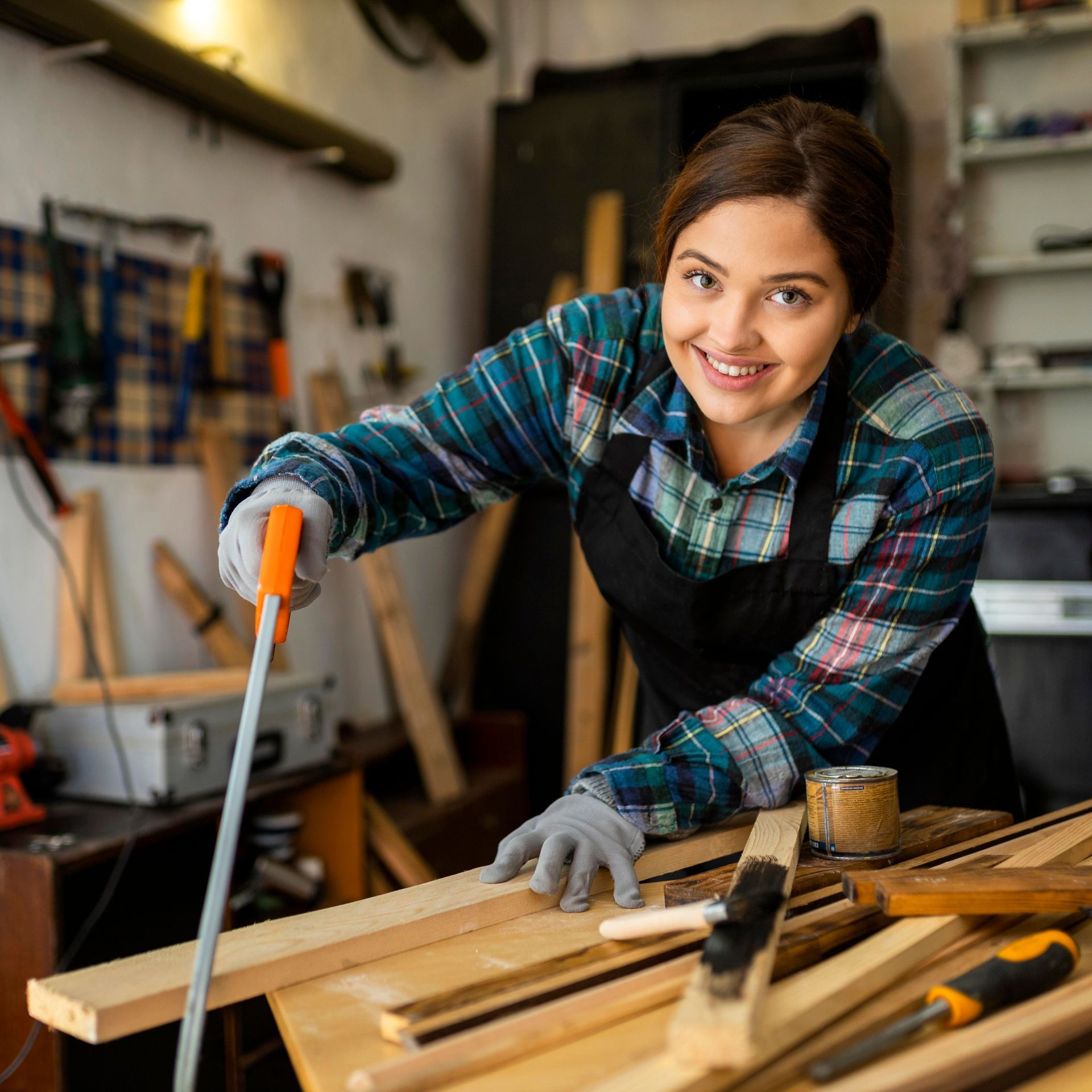 Woman in workshop using a hand saw on wood, wearing safety gloves and apron, smiling.