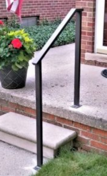 Black metal handrail on a concrete step leading up to a brick building.