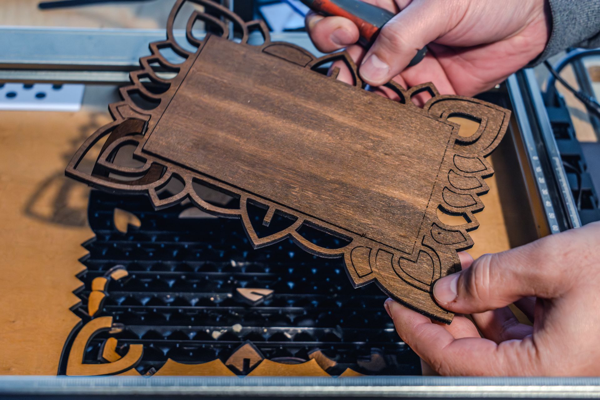 Hands holding a laser-cut, stained wood design. A laser cutter and work surface are in the background.
