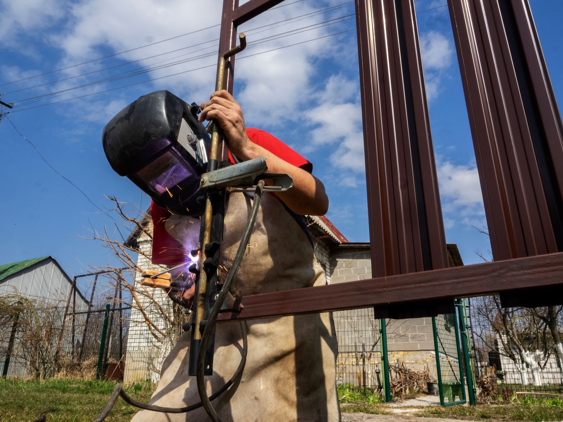 Welder in protective gear welding a brown metal fence outdoors on a sunny day.