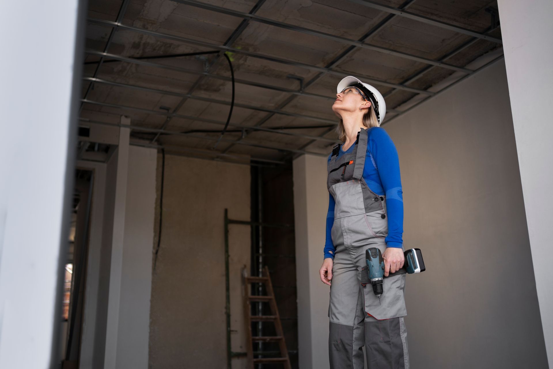 Construction worker in overalls and hard hat inspects unfinished ceiling, holding a drill.