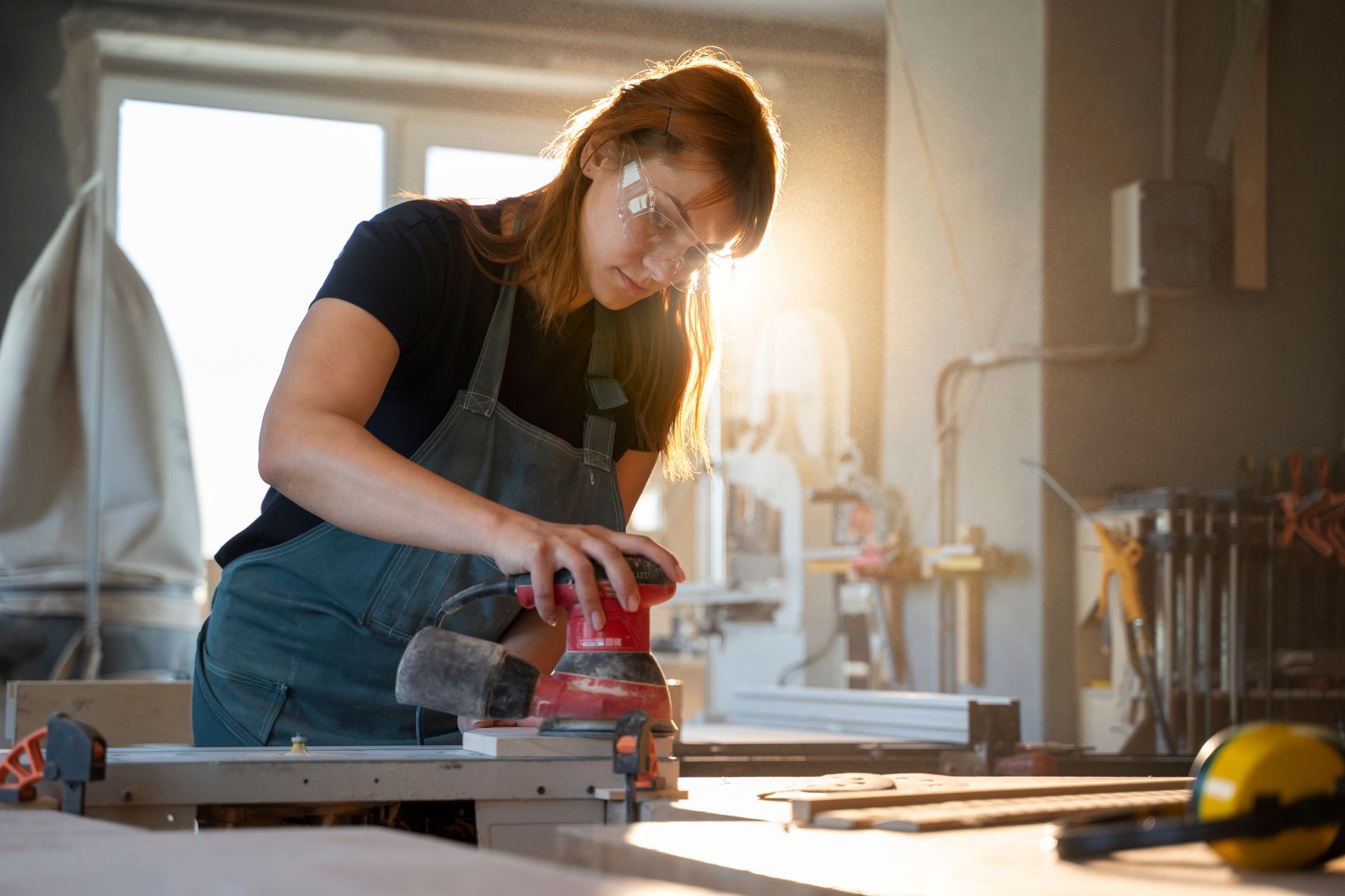 Woman sanding wood with a power sander in a workshop, wearing safety glasses and apron.