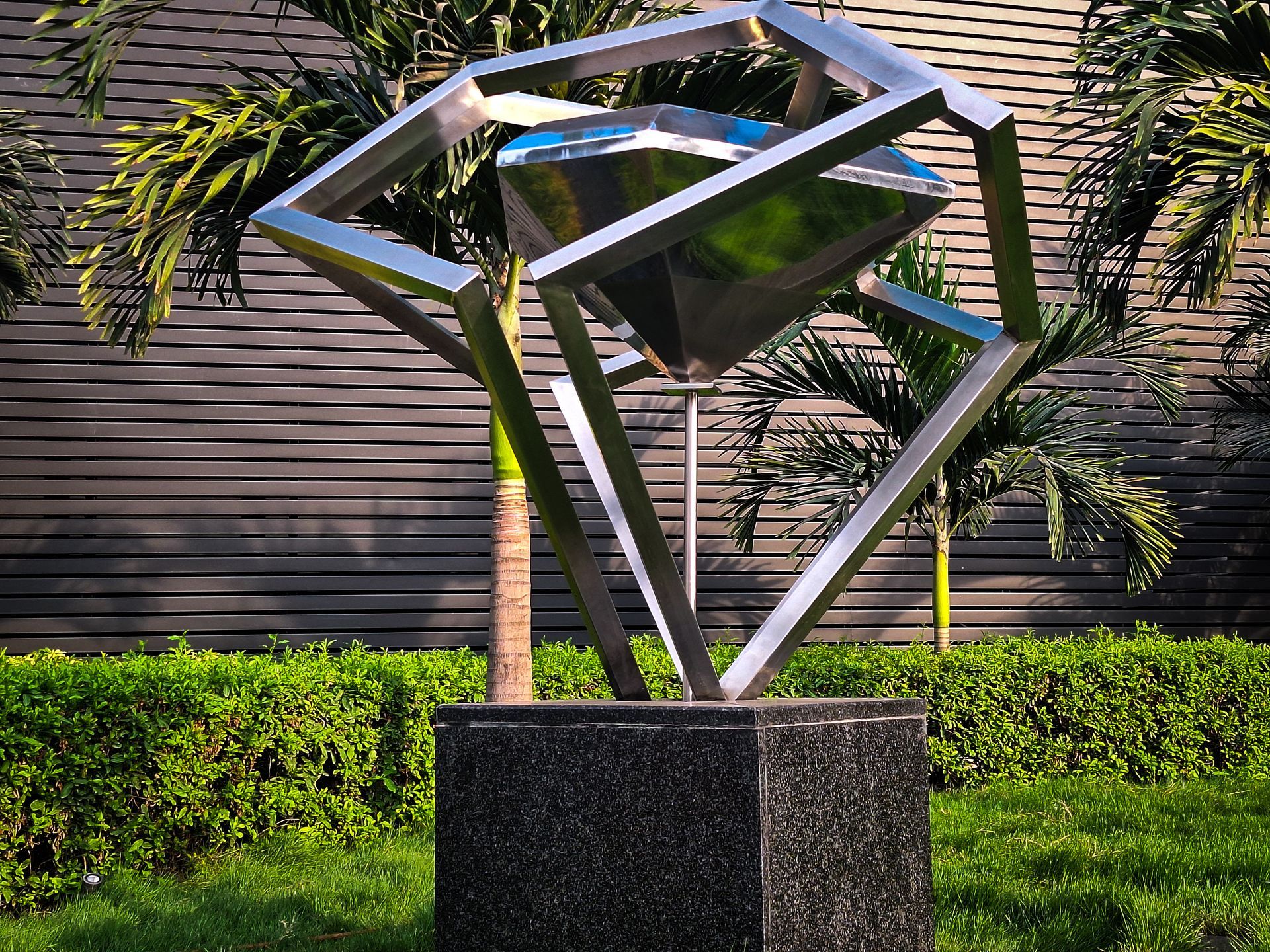 Sculpture: a geometric diamond shape encased in a metal frame, set on a dark granite base in a garden.