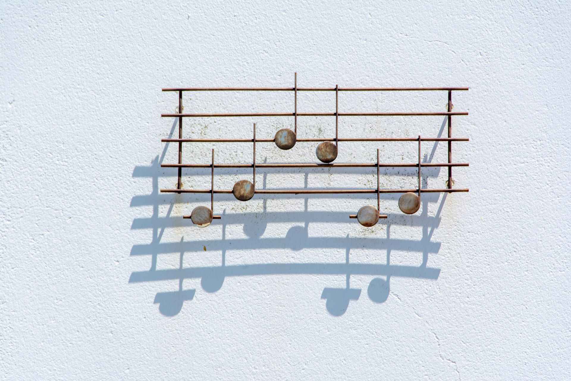 Rusty metal music staff with spherical notes mounted on a white wall, casting a shadow.