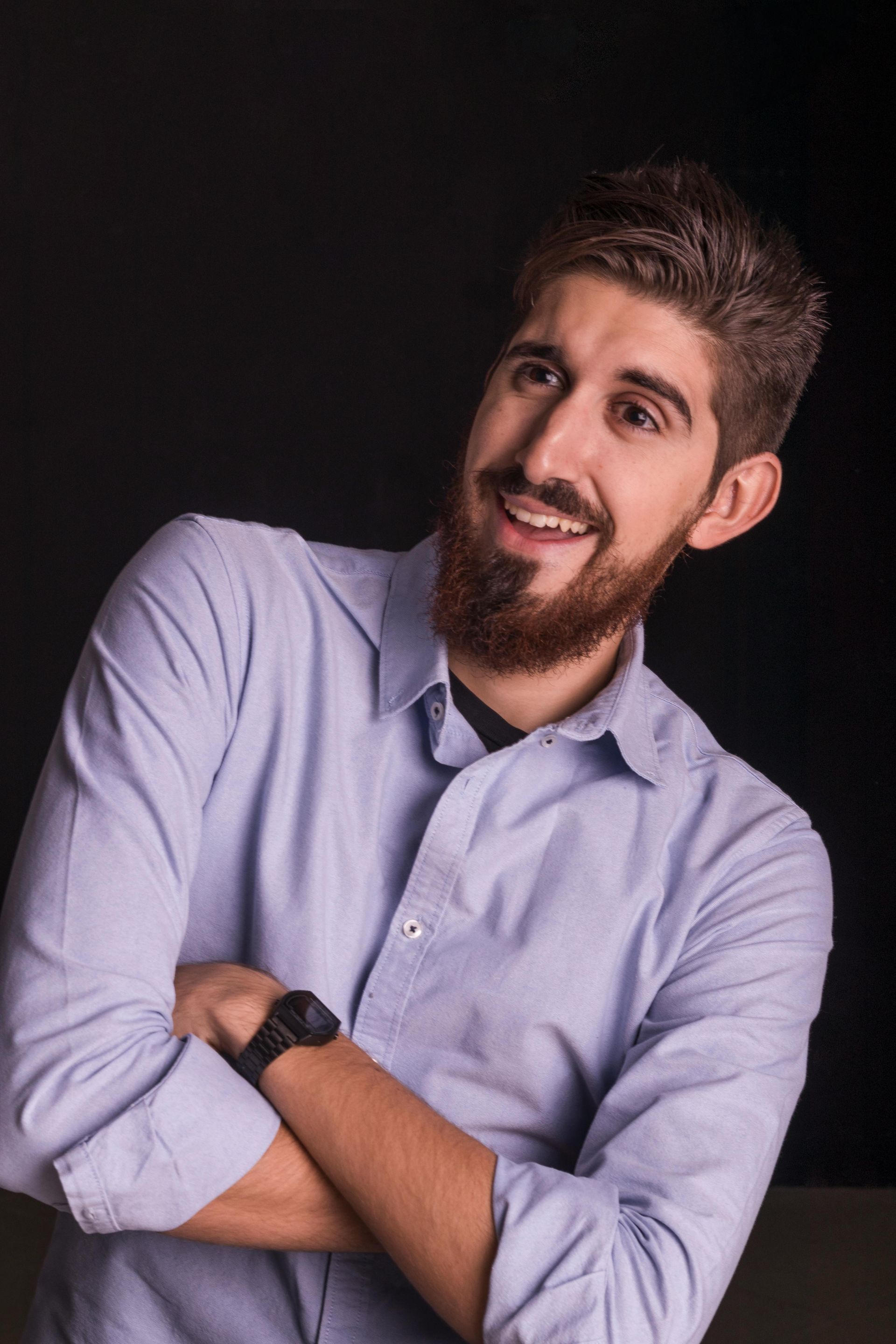 Man with beard and light blue shirt smiles, arms crossed, looking upwards. Dark background.
