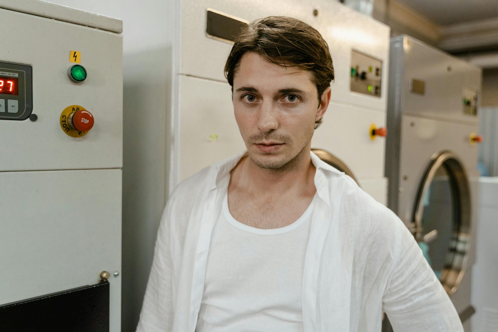 Man in white shirt and undershirt poses in a laundromat, near machinery.