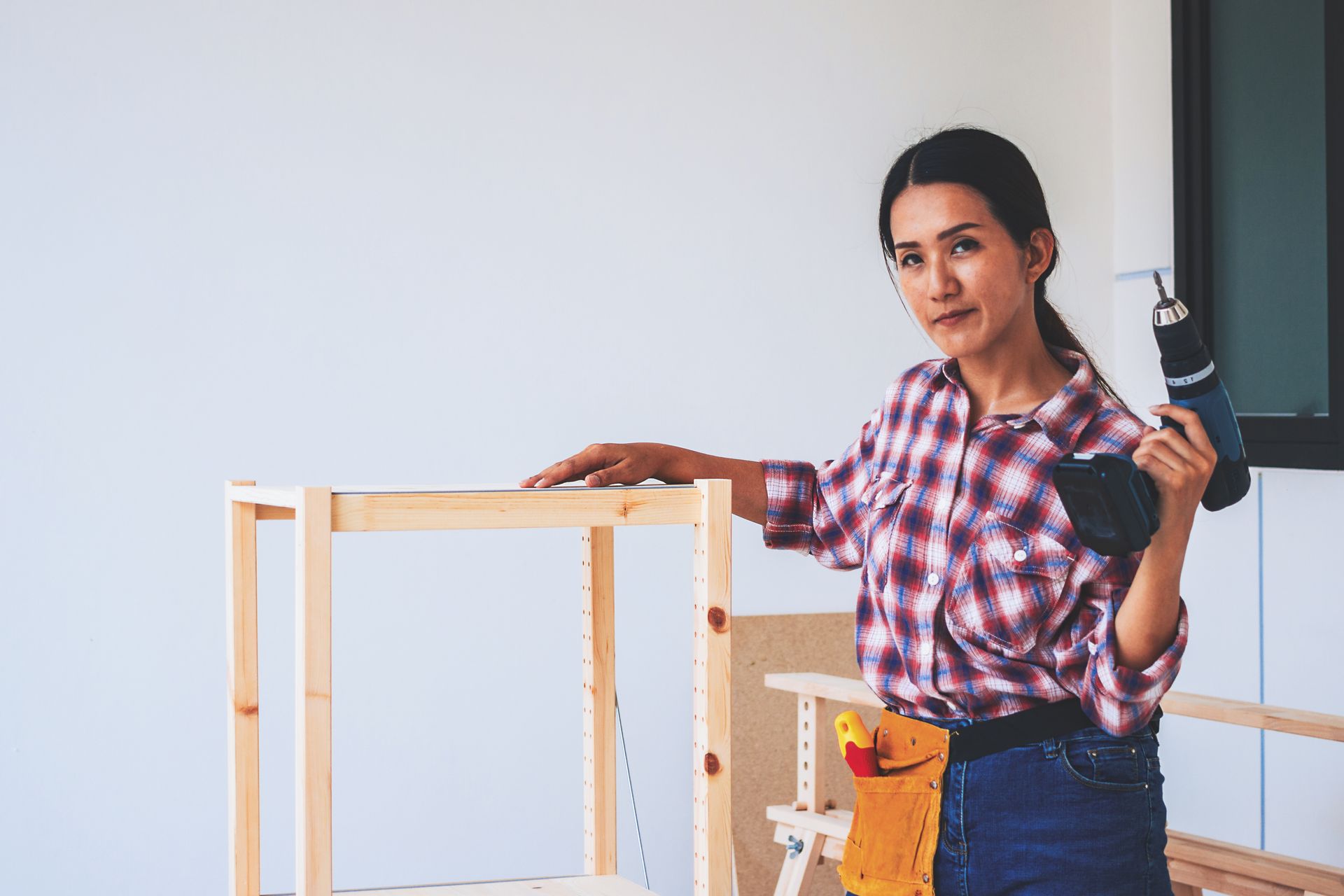 Woman assembling wooden shelf with a drill in a room with white walls.