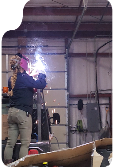 Woman welding in a workshop, wearing protective gear. Sparks fly from the metal.