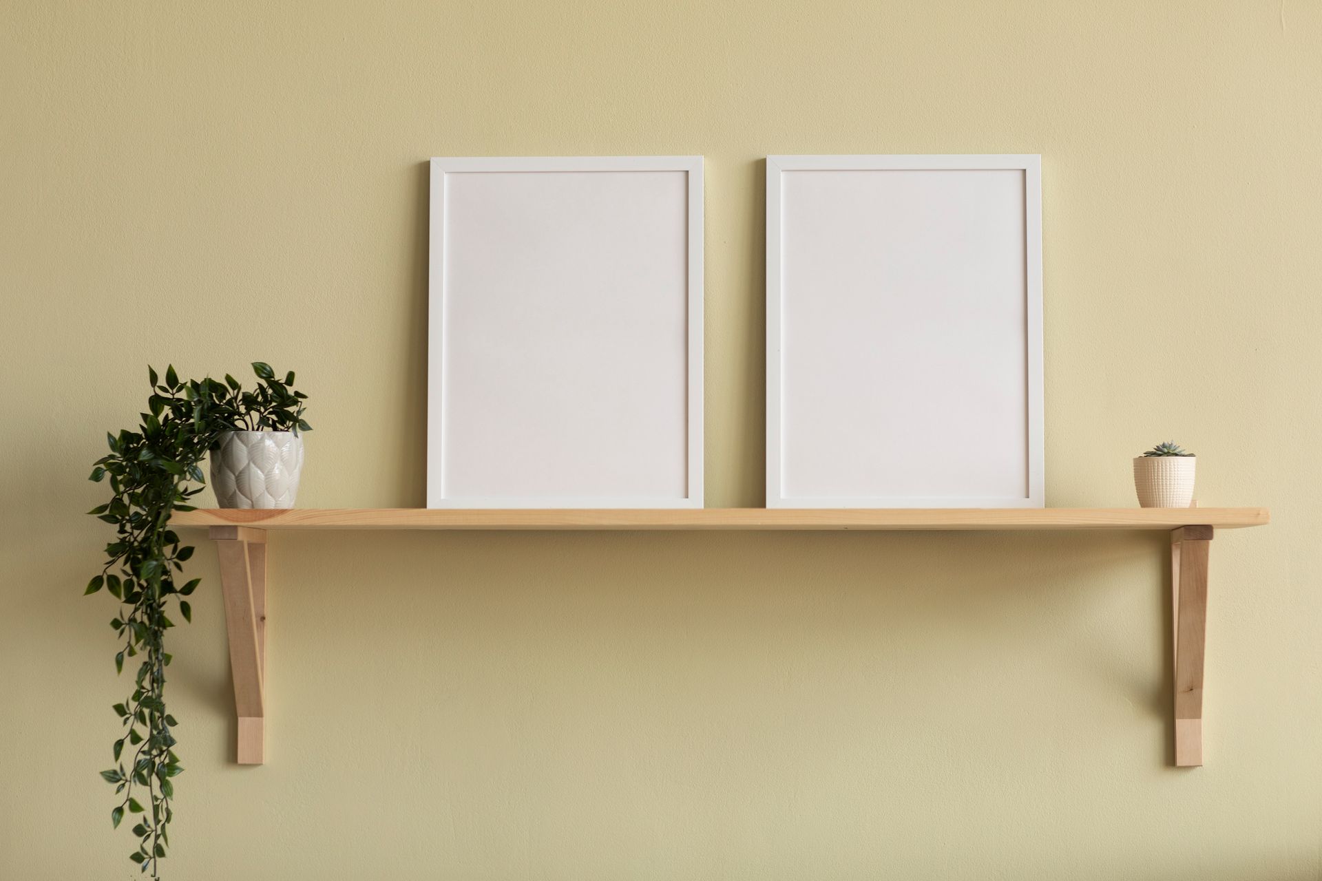 Shelf with two white framed blank pictures, two small plants, against a light yellow wall.