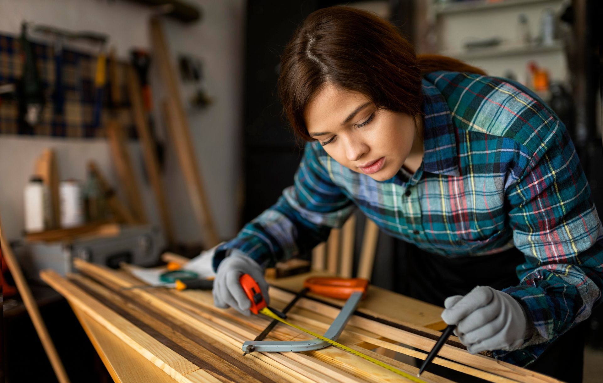 Woman in plaid shirt using a measuring tool on a wood plank in a workshop.