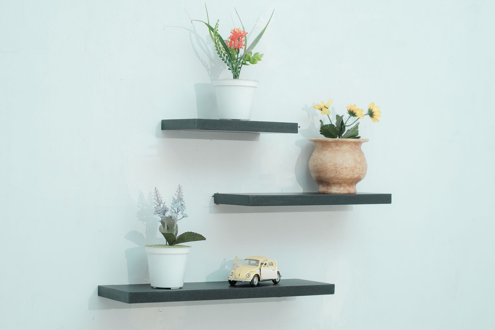 Three gray floating shelves on a white wall; each holds a potted plant and a toy car.