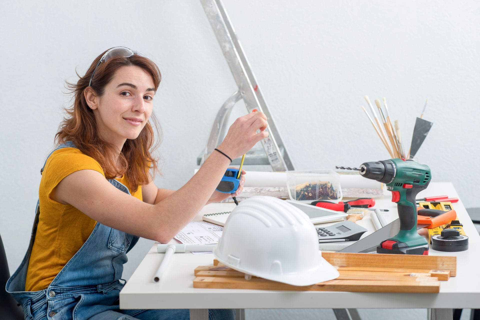 Woman in overalls smiles, holding a tape measure at a work table with tools, a ladder in background.
