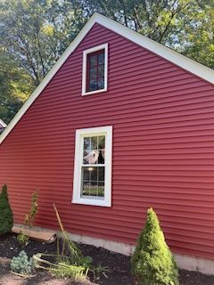 Red-sided building with white trim and windows, set against trees and green foliage.