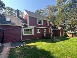 Red house with white trim and green lawn, surrounded by trees under a blue sky.