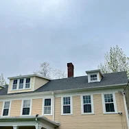 Tan house with multiple windows, dormers, and a brick chimney under a cloudy sky.
